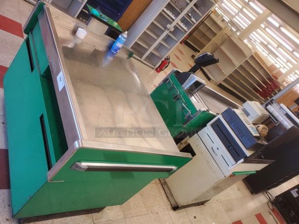 Green supermarket checkout counter with a stainless steel top, featuring storage compartments. Slightly worn but functional.