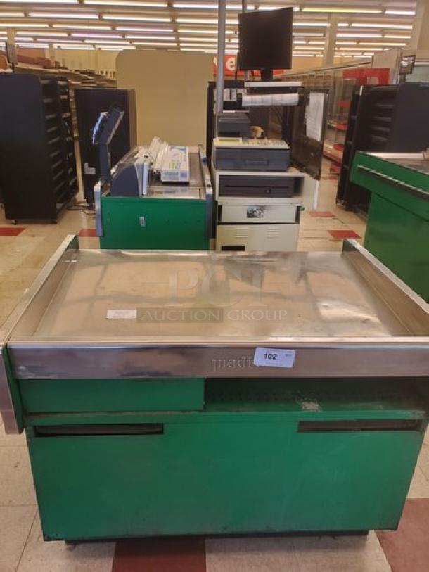 Supermarket checkout counter with green base, stainless steel top, and attached conveyor belt, showing some wear.