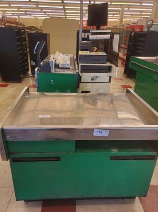 Supermarket checkout counter with green base, stainless steel top, and attached conveyor belt, showing some wear.