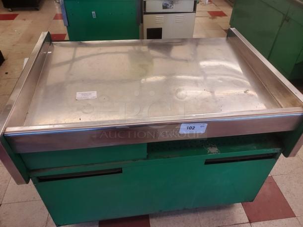 Stainless steel supermarket checkout counter with green base, moderate wear, inventory tag #102 visible.