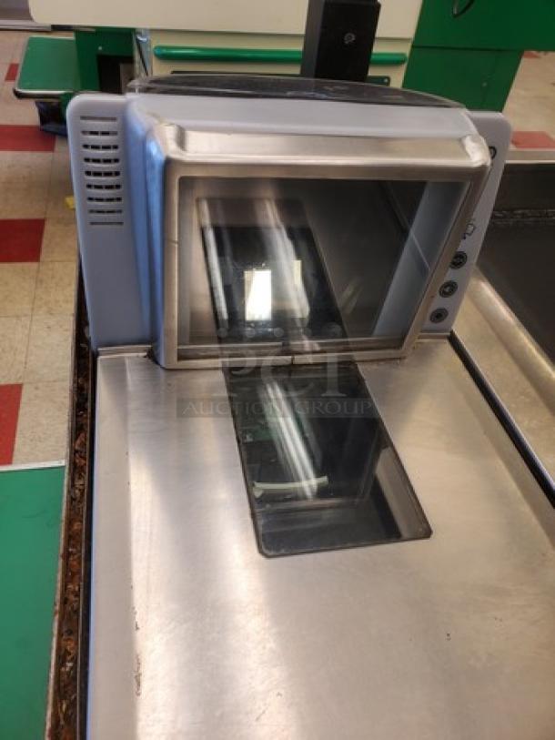 Supermarket checkout counter with scanner, metallic surface, and integrated digital display. Used condition, minor wear visible.