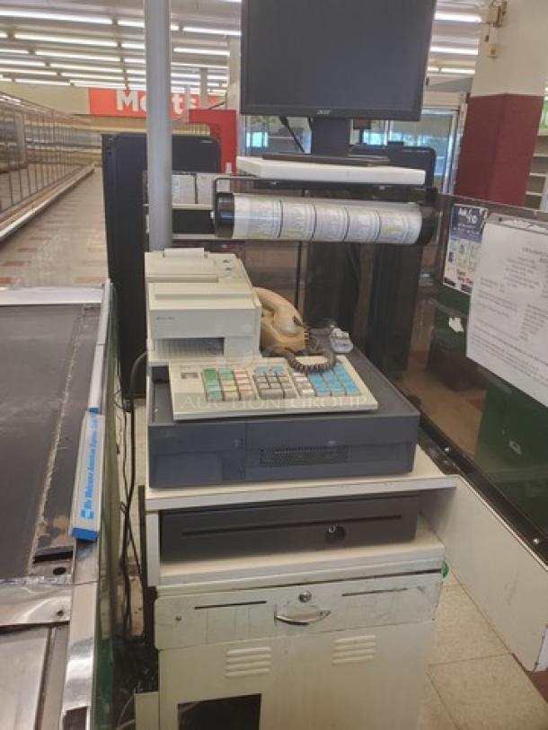 Supermarket checkout counter with built-in cash register, receipt printer, and monitor; shows visible wear.