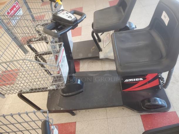 Motorized shopping cart, Amigo brand, with a metal basket and padded seat. Features steering controls and sturdy platform.