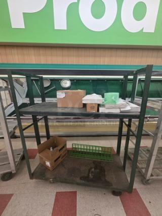 Industrial shelf cart with two metal shelves, green frame, used condition. Includes various stored items. Movable on wheels.