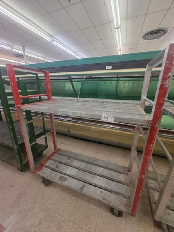 Aluminum mobile shelf cart with two tiers and red side accents, showing signs of wear. Mounted on wheels for easy movement.
