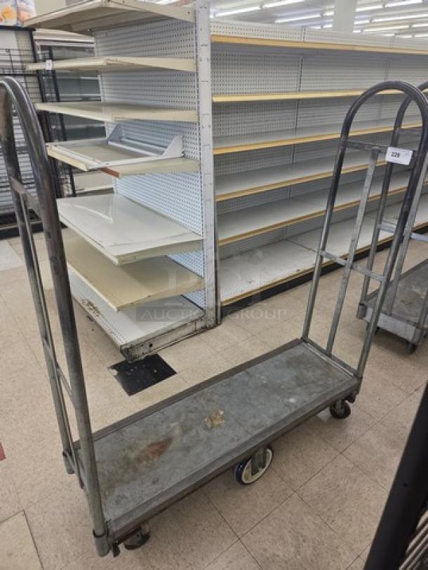 Metal stock cart with two curved handles, four wheels, and a worn surface. Used condition, ideal for warehouse or retail.