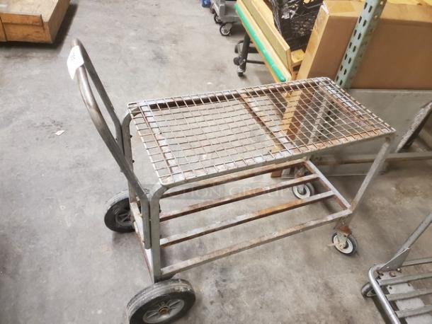 A metal retail dolly cart with a grid top, four wheels, and a push handle. Shows signs of wear, suitable for warehouse use.