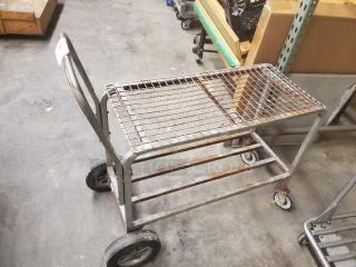 A metal retail dolly cart with a grid top, four wheels, and a push handle. Shows signs of wear, suitable for warehouse use.