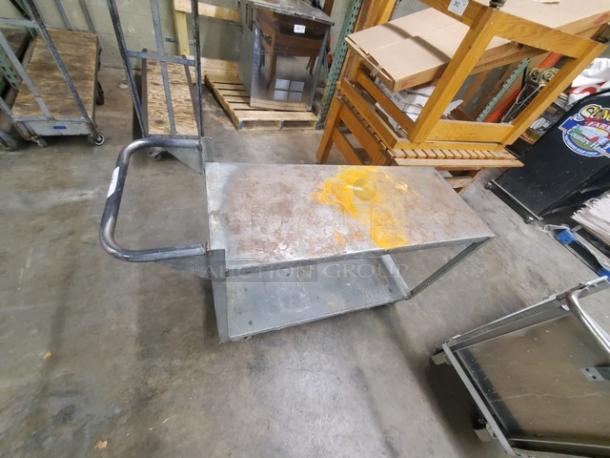Metal utility cart with two shelves and a push handle, showing wear and rust stains, suitable for warehouse use.