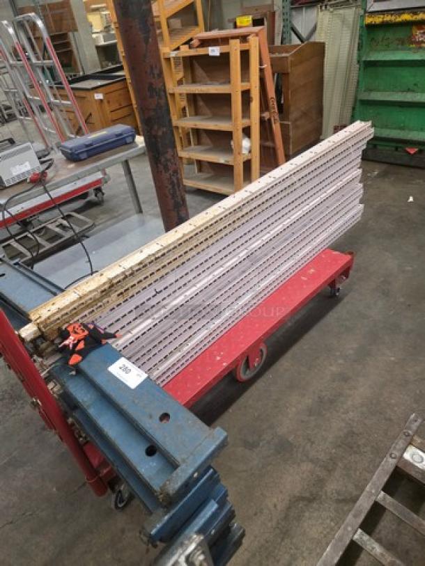 Lot of pallet rack beams stacked on a red dolly in a warehouse. Metal beams show markings and wear, indicating heavy use.