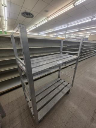 Aluminum mobile shelf cart with three tiers, industrial design, in used condition. Markings visible on middle shelf.