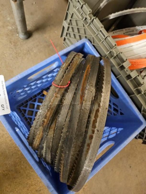 Lot of band saw blades, assorted sizes, and lengths, showing some rust and use wear, bundled with a red tie, in a blue crate.