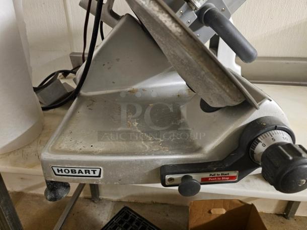 Hobart slicer with manual controls, visible wear, and brand label. Features adjustable thickness dial, stainless steel body.