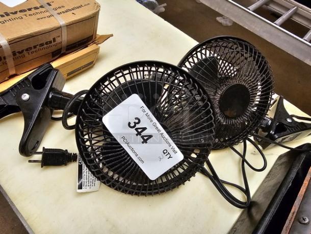 Two small black clip-on fans with mesh guards and power cords, in used condition. Auction tag marked "344" visible.