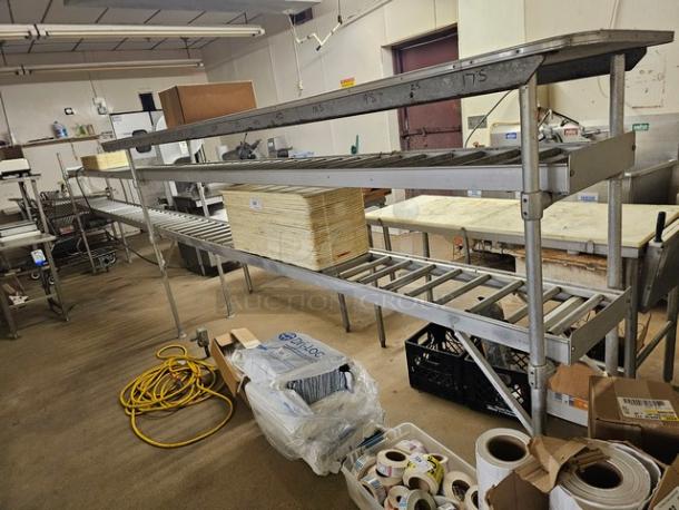 Stainless steel roller conveyor system in industrial setting, features rollers and upper shelving, used condition.