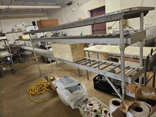 Stainless steel roller conveyor system in industrial setting, features rollers and upper shelving, used condition.