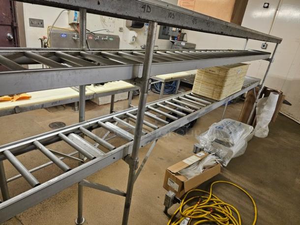 Stainless steel roller conveyor system with two levels, used condition. Includes wood sheets and various tools beneath.