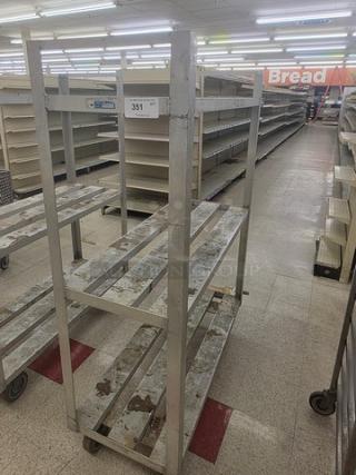 Aluminum mobile shelf cart with four tiers and caster wheels, showing signs of wear. Ideal for storage or moving items.