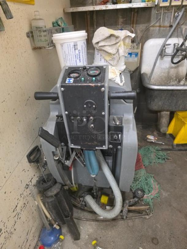 Tennant floor scrubber in used condition with visible control panel and hoses, stored in a maintenance area.