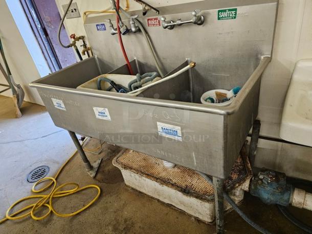 Stainless steel 3 compartment sink with labeled wash, rinse, sanitize faucets. Used condition, includes plumbing fixtures and hoses.