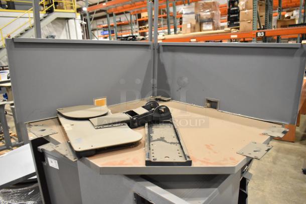 Gray L-shaped desk setup in industrial setting, featuring a heavy-duty monitor arm and partition panels. Some wear visible.