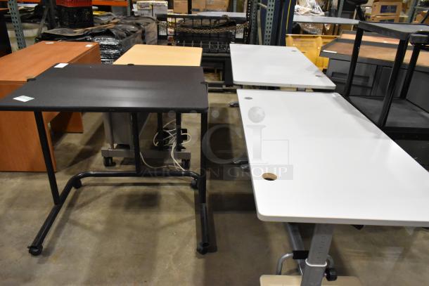 Four office desks/tables, varied styles and colors, on display in warehouse. Includes white, black, wood finishes.