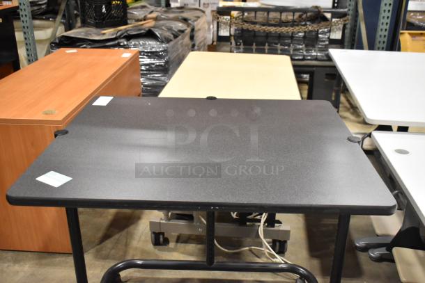 Four various desks/tables including a black, wood, and beige top. Good condition. Cables and labels visible.