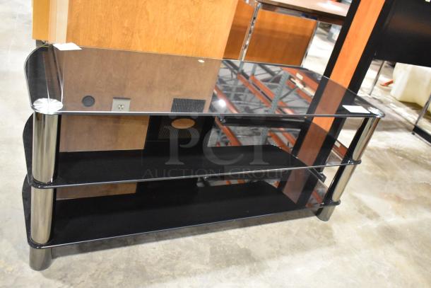Black television stand with glass top, two under shelves, and silver metal supports. Appears in excellent condition.
