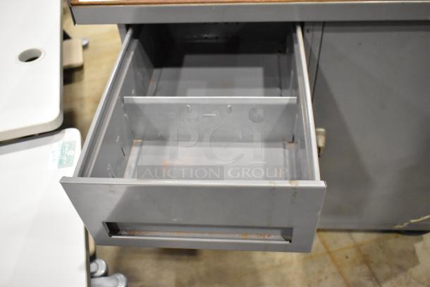 Gray metal cart with four drawers and a door on commercial casters. Drawers shown open, minor wear visible.