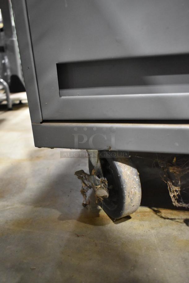 Gray metal cart with four drawers and a door on commercial casters, showing wear and debris on wheels.