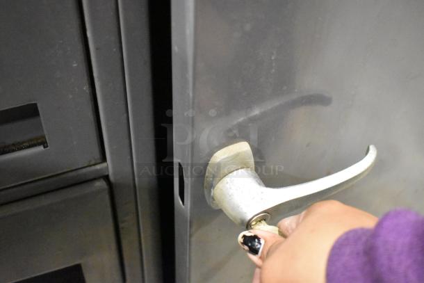 Gray metal cart with four drawers, a lockable door, and commercial casters. Shown in used condition with hand on handle.