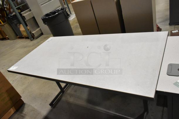 Rectangular office desk with a light gray tabletop and black metal legs in a warehouse setting. Slight wear visible.