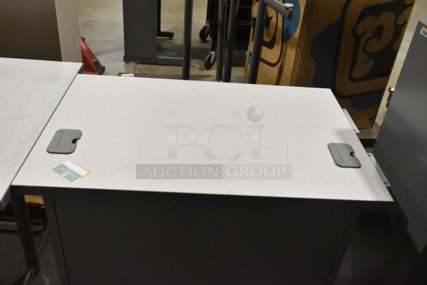 Three office items including a grey desk with visible cable ports, pristine condition. Label indicates university origin.