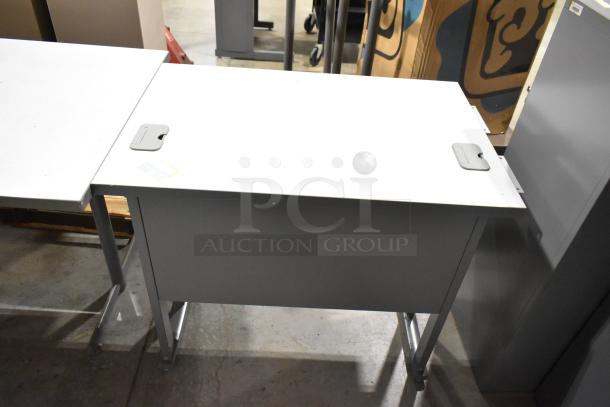 Gray desk with metal legs and white top; visible cable management cutouts, part of a lot with bookshelf and another desk.