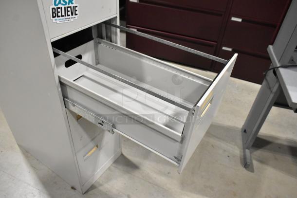 Gray metal 4-drawer filing cabinet, open top drawer, sticker reading "Believe," sturdy condition, office setting.