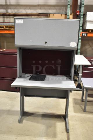 Gray desk with overhead cabinet, sturdy metal frame, in good condition. Features a lockable cabinet door and a flat workspace.