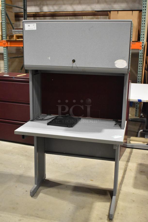 Gray desk with overhead cabinet, sturdy design, minor wear. Includes compartment for cable management. Auction label present.