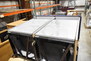 2 gray double trash can shells on casters, industrial setting, durable construction, moderate wear, useful for easy waste management.