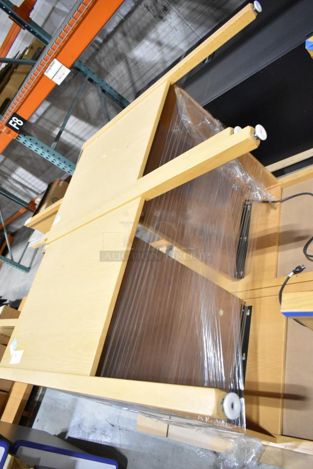 Two wood pattern desks stacked, wrapped in plastic. Light wood finish, metal brackets, and visible power cords. Good condition.