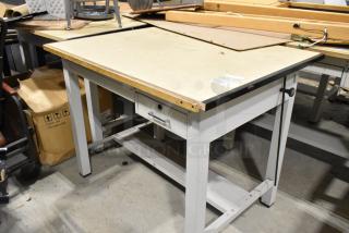 Gray metal table with wooden tabletop, featuring a drawer and sturdy legs. Slight wear visible, suitable for workshop use.