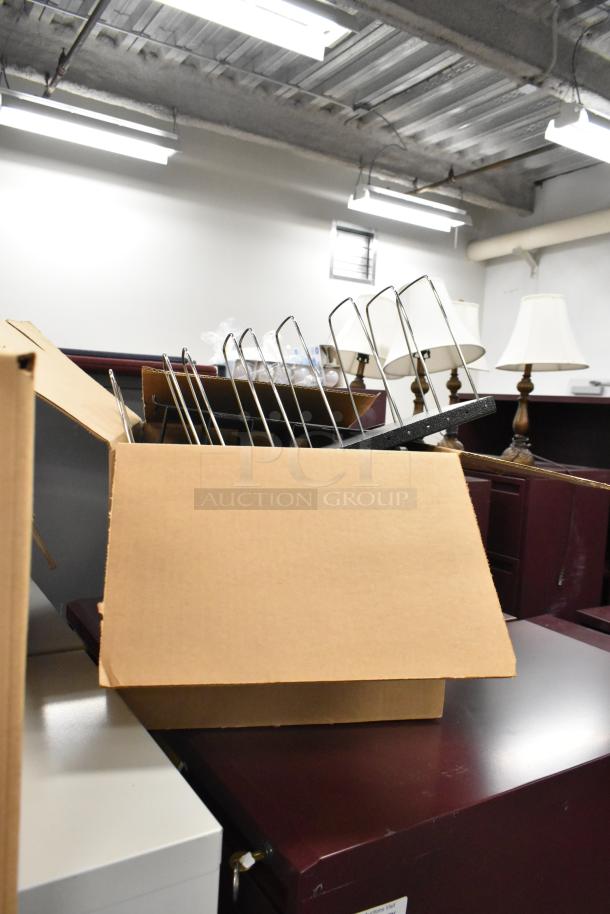 Cardboard box with office items, including a metal rack and staplers, on a wooden surface. Background lamps visible.