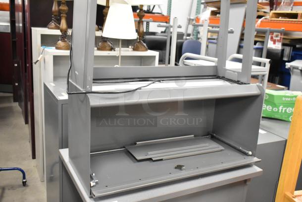 Gray desk with overhead cabinet, metal construction, minor wear, sturdy frame, suitable for office use, power cables attached.