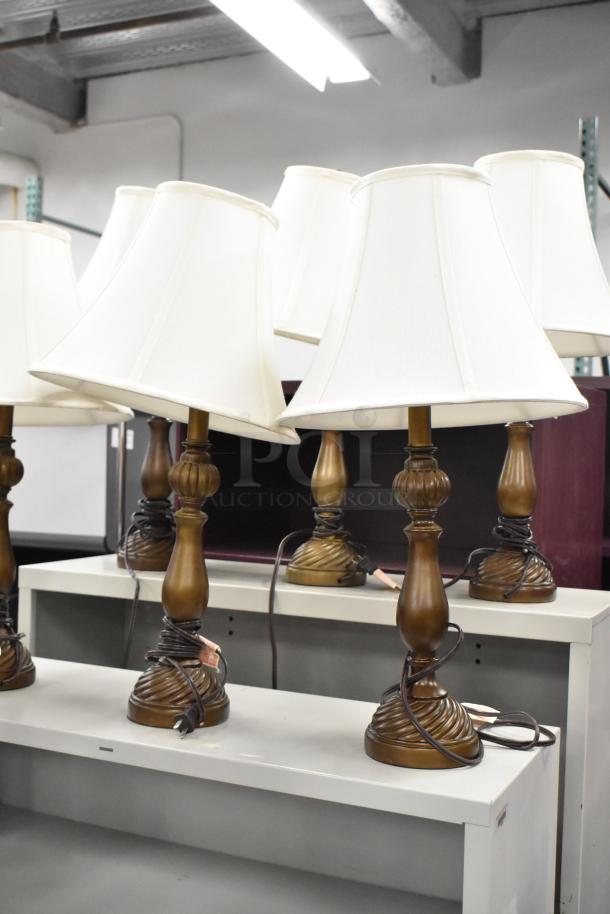Eight bronze table lamps with white fabric shades, displaying intricate bases. Each lamp is in good condition.