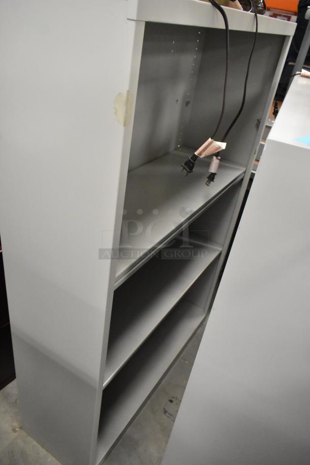 Two gray bookshelves with three shelves each, visible scuffing, and attached electrical cords. Used condition.