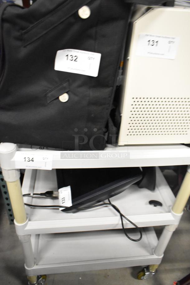 3-tier cart on casters, white shelves, labeled "134", holds electronic equipment and a black jacket, in used condition.