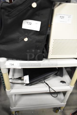 3-tier cart on casters, white shelves, labeled "134", holds electronic equipment and a black jacket, in used condition.