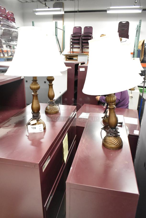 Four table lamps with white shades and ornate bronze bases, displayed on purple filing cabinets in used condition.