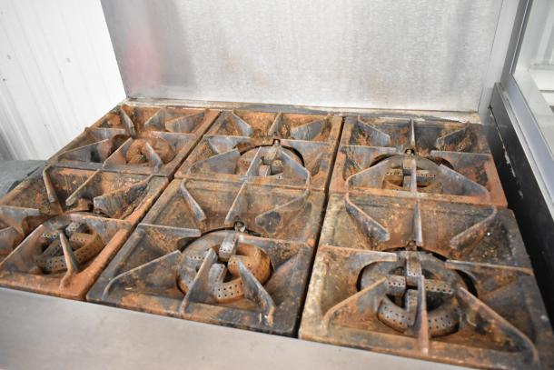 Stainless steel commercial gas 6 burner range by Cooking Performance Group, with visible rust and wear on burners. Features backsplash and overshelf.