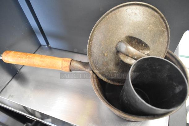 Metal saucepan with wooden handle, shows signs of wear and rust. Lid and black cylindrical container included.