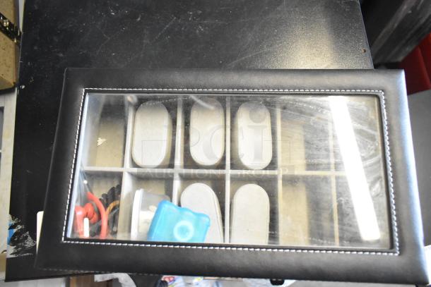 Watch case with glass top, black leather exterior, visible compartments, in good condition, includes items like cords and a small container.